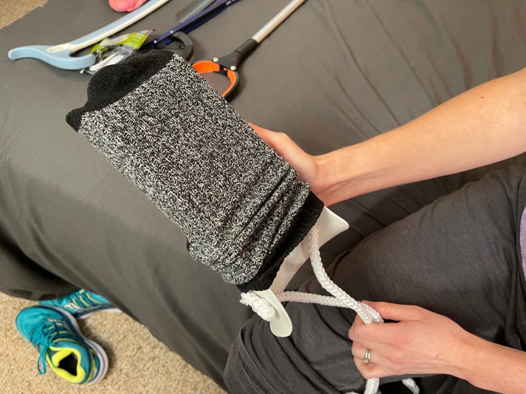 A person demonstrates using a sock aid device by pulling a black and gray sock onto the aid's curved frame. The white ropes attached to the device are held firmly. Lower Body Dressing After Hip Replacement Surgery