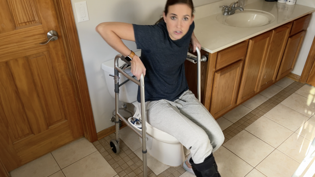 Person using the walker as support to safely lower themselves onto the toilet while maintaining a non-weight-bearing position on the injured leg. How to Use Toilet When Non-Weight Bearing with Crutches or Walker - EquipMeOT