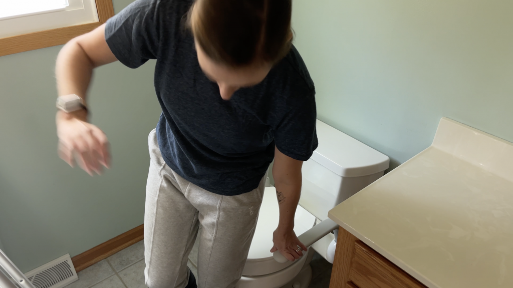 A person stabilizes themselves using toilet safety rails before lowering onto a toilet seat. The individual is wearing light gray sweatpants and a navy blue shirt, with one hand gripping the rail and the other extended for balance. How to Use Toilet When Non-Weight Bearing with Crutches or Walker - EquipMeOT