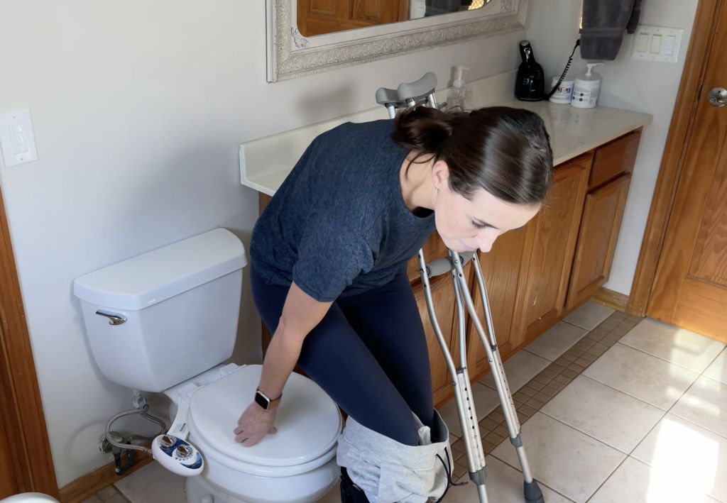 Person using crutches in a bathroom, holding onto a counter and the toilet seat for support while lowering themselves onto the toilet. They are wearing a dark blue shirt and managing their pants to sit safely while maintaining balance. How to Use Toilet When Non-Weight Bearing with Crutches or Walker - EquipMeOT