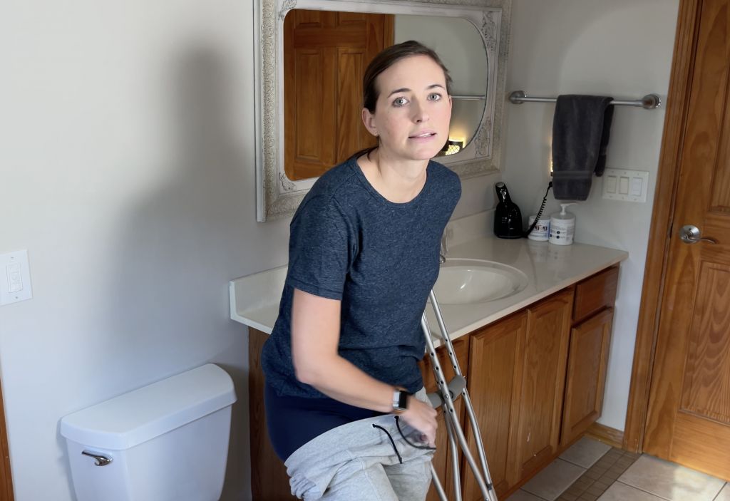 Person using crutches in a bathroom, holding onto a solid surface for balance while managing their clothes. They are wearing a dark blue shirt and light gray pants, preparing to use the toilet while non-weight-bearing. How to Use Toilet When Non-Weight Bearing with Crutches or Walker - EquipMeOt