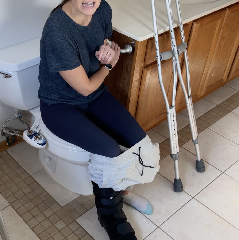 Person sitting on a toilet in a bathroom, wearing a medical boot on their non-weight-bearing leg, which is elevated slightly or resting lightly on the floor. They are using their hands for balance while their crutches lean against the nearby counter. How to Use Toilet When Non-Weight Bearing with Crutches or Walker - EquipMeOT