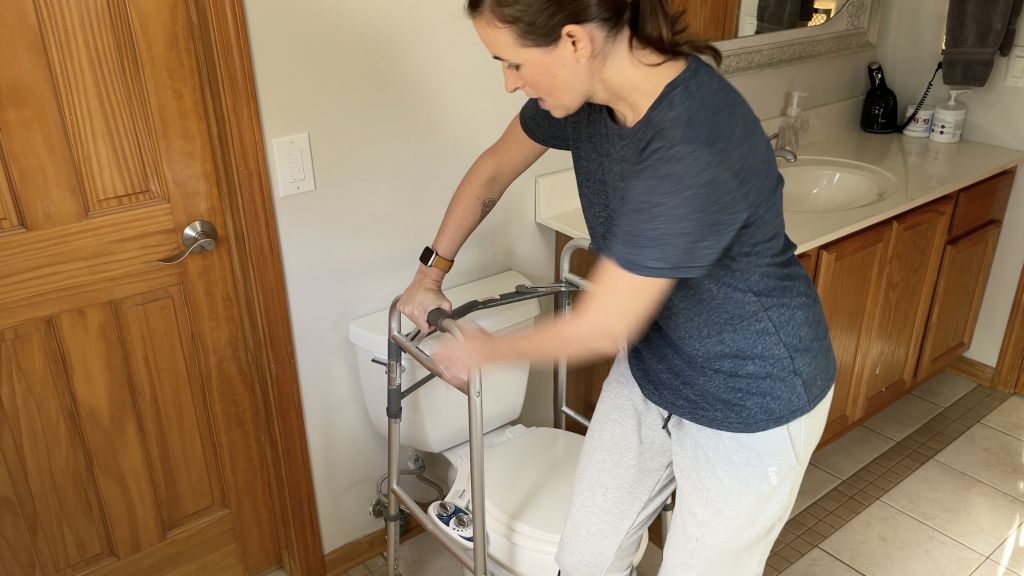 Individual pivoting one hand at a time on a walker placed in front of a toilet, demonstrating a careful technique to ensure stability and safety while managing a non-weight-bearing leg. How to Use Toilet When Non-Weight Bearing with Crutches or Walker - EquipMeOT