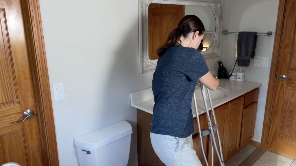 Person in a bathroom placing crutches in a stable position near a countertop for balance. They are wearing a dark blue shirt and light gray pants, preparing to safely transition to using the toilet while being non-weight-bearing. How to Use Toilet When Non-Weight Bearing with Crutches or Walker - EquipMeOt