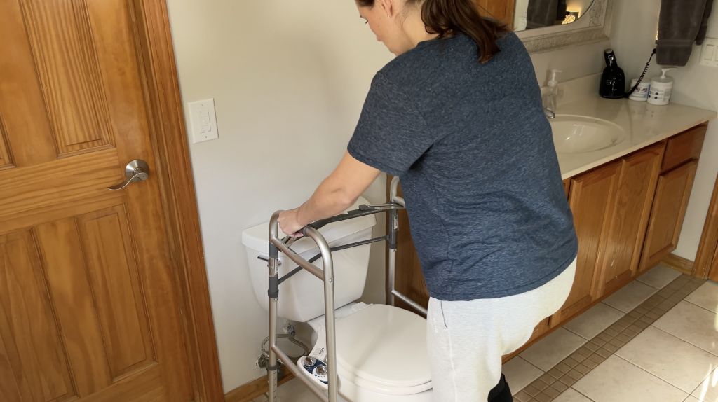 Person positioning a walker in front of a toilet tank in a bathroom, preparing to use it for support while navigating the toilet. The individual is wearing a medical boot on their non-weight-bearing leg. How to Use Toilet When Non-Weight Bearing with Crutches or Walker - EquipMeOT