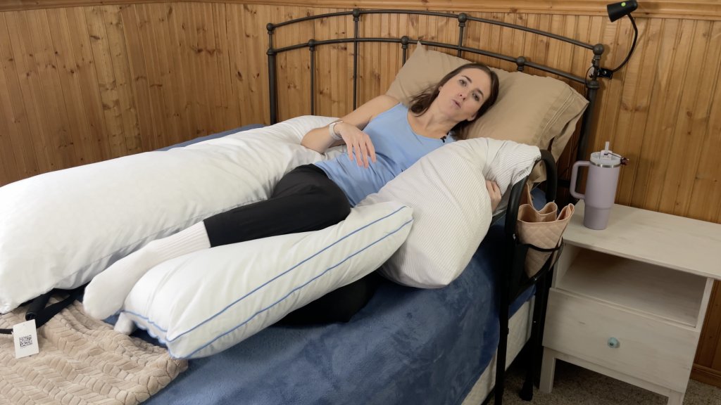 A woman is turning in bed using the log roll technique, supported by multiple pillows. Her knees are bent in a right angle and her back is straight. She is wearing a light blue tank top. How to Sleep After a C-Section - EquipMeOT