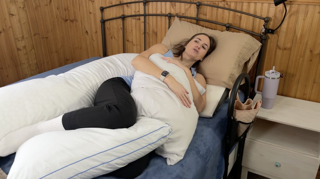 A woman is lying on her side in bed, supported by multiple pillows. She is wearing a light blue tank top, black pants, and white socks. Her legs are cushioned by large white pillows, while she hugs another pillow against her chest. She is resting forward but remains by a body pillow behind her back.