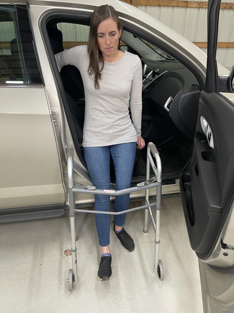 A woman gets into a car using a walker for support. She holds onto the car's door frame with one hand and steadies herself on the seat with the other. She is facing away from the car. Dressed in a light gray long-sleeve shirt, blue jeans, and black slip-on shoes, she demonstrates a safe method for enter a vehicle with mobility assistance. How to Get In and Out of the Car After Hip Replacement