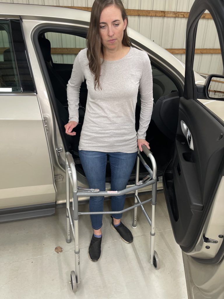 A woman stands outside a car using a walker for support. She has exited the vehicle and steadies herself with both hands on the walker. She is wearing a light gray long-sleeve shirt, blue jeans, and black slip-on shoes. The car door is open, revealing part of the interior, illustrating the use of a mobility aid for a safe and stable transition. How to Get In and Out of the Car After Hip Replacement