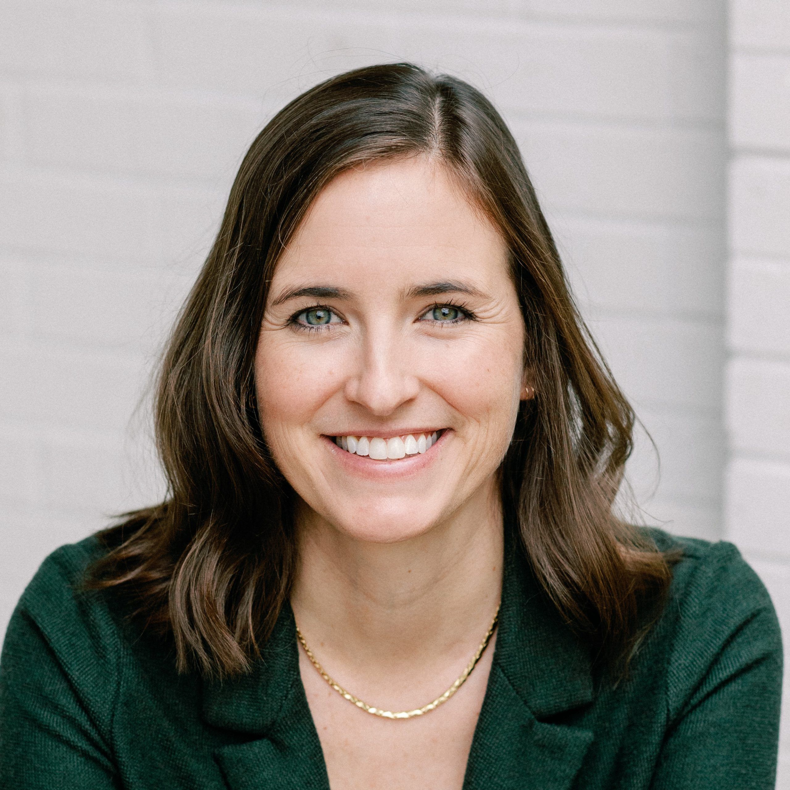 A smiling woman seated outdoors, wearing a dark green blazer over. She has shoulder-length brown hair and is accessorized with a delicate gold chain necklace and a wristwatch. The background features a light-colored brick wall, creating a clean and professional portrait setting.